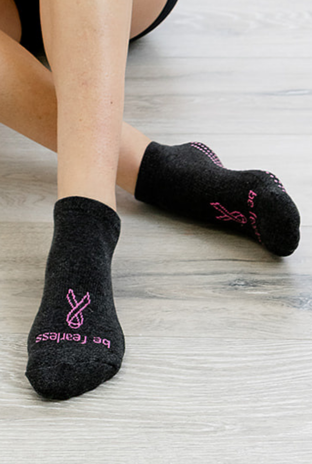 A person wearing black grip socks with pink dots and text reading 'BE BRAVE' and 'FEARLESS' seated and holding a pink exercise ring.